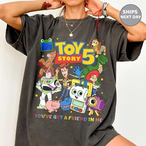 May include: A charcoal gray t-shirt featuring a colorful graphic print of Toy Story characters including Woody, Buzz Lightyear, Rex, and Jessie. The design includes the text "TOY STORY 5" in large yellow and blue letters and "YOU'VE GOT A FRIEND IN ME" below.