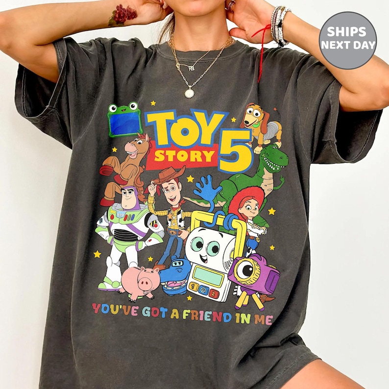May include: A charcoal gray t-shirt featuring a colorful graphic print of Toy Story characters including Woody, Buzz Lightyear, Rex, and Jessie. The design includes the text "TOY STORY 5" in large yellow and blue letters and "YOU'VE GOT A FRIEND IN ME" below.