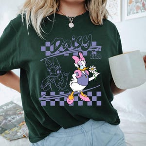May include: A dark green short-sleeve t-shirt featuring a graphic of Daisy Duck holding a white daisy flower. The design includes a purple checkered background, the text Daisy, and the year 1928 in a stylized font, with a line art outline of Daisy.