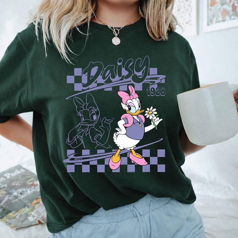 May include: A dark green short-sleeve t-shirt featuring a graphic of Daisy Duck holding a white daisy flower. The design includes a purple checkered background, the text Daisy, and the year 1928 in a stylized font, with a line art outline of Daisy.