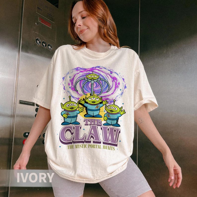 May include: An ivory short-sleeve t-shirt featuring a graphic of three green three-eyed aliens under a mechanical claw and a purple swirling portal. The text reads THE CLAW and THE MYSTIC PORTAL AWAITS in a bold, stylized purple font.