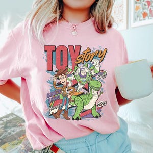 May include: Pink t-shirt featuring a colorful graphic of Toy Story characters Woody, Buzz Lightyear, and Rex. The design includes the text "TOY STORY" and "Since 1995". The shirt is a crew neck style.
