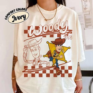 K&ouml;nnte beinhalten: Ein elfenbeinfarbenes T-Shirt mit einem Grafikdruck der Figur Woody aus Toy Story. Das Design umfasst eine Strichzeichnung von Woody, eine farbige Illustration von Woody vor einem gelben Sheriffstern, ein Karomuster und den Text Woody 199S.