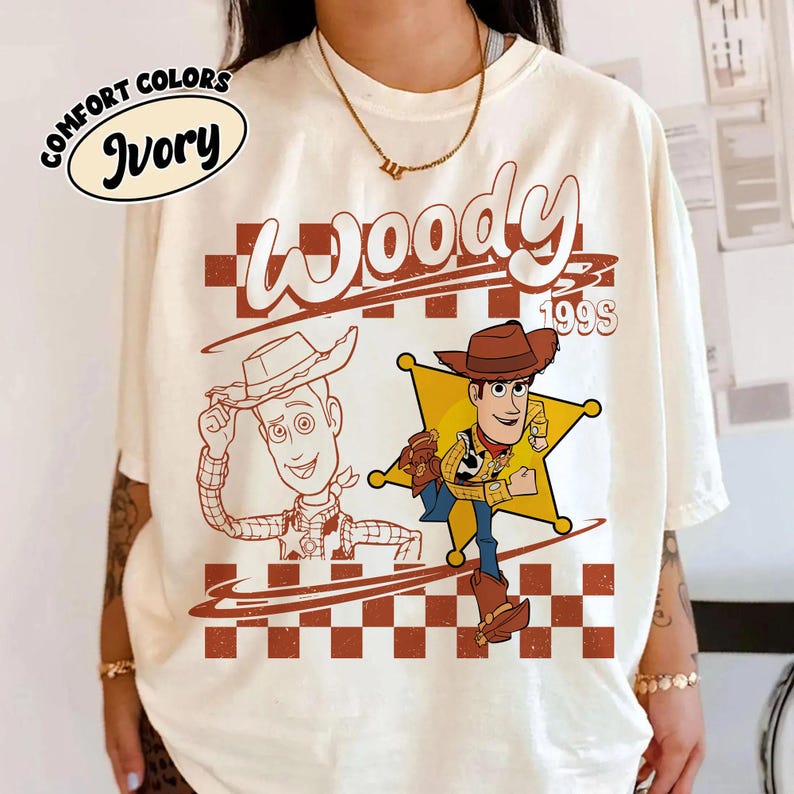 K&ouml;nnte beinhalten: Ein elfenbeinfarbenes T-Shirt mit einem Grafikdruck der Figur Woody aus Toy Story. Das Design umfasst eine Strichzeichnung von Woody, eine farbige Illustration von Woody vor einem gelben Sheriffstern, ein Karomuster und den Text Woody 199S.