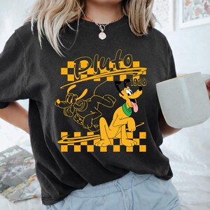 May include: A charcoal gray short-sleeve t-shirt featuring a yellow and black checkered graphic with the character Pluto, the text Pluto, and the year 1928. The design includes a yellow outline of the character and a solid yellow Pluto figure.