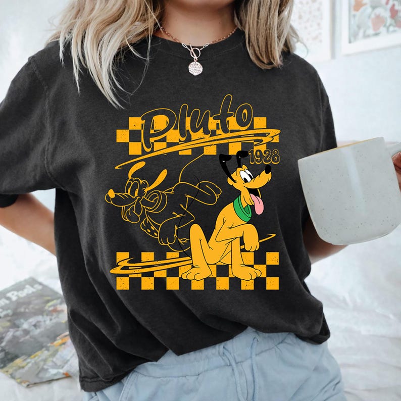 May include: A charcoal gray short-sleeve t-shirt featuring a yellow and black checkered graphic with the character Pluto, the text Pluto, and the year 1928. The design includes a yellow outline of the character and a solid yellow Pluto figure.