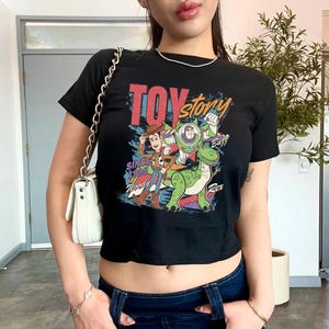 May include: Black crop top featuring a colorful graphic of Toy Story characters, including Woody, Buzz Lightyear, and Rex. The text "TOY STORY" and "SINCE 1995" are also displayed. The shirt has short sleeves and a round neckline.