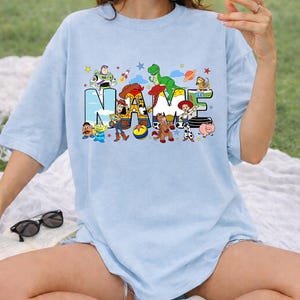May include: Light blue t-shirt featuring the word "NAME" in a colorful, cartoon-style font. The letters are decorated with characters from a popular animated movie. The shirt is a relaxed fit and has short sleeves.