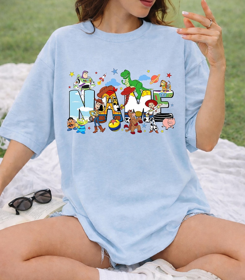 May include: Light blue t-shirt featuring the word "NAME" in a colorful, cartoon-style font. The letters are decorated with characters from a popular animated movie. The shirt is a relaxed fit and has short sleeves.