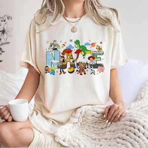 May include: Cream-colored t-shirt featuring the word "NAME" in a colorful, cartoonish font. The letters are decorated with characters from a popular animated movie. The shirt has short sleeves and a relaxed fit.