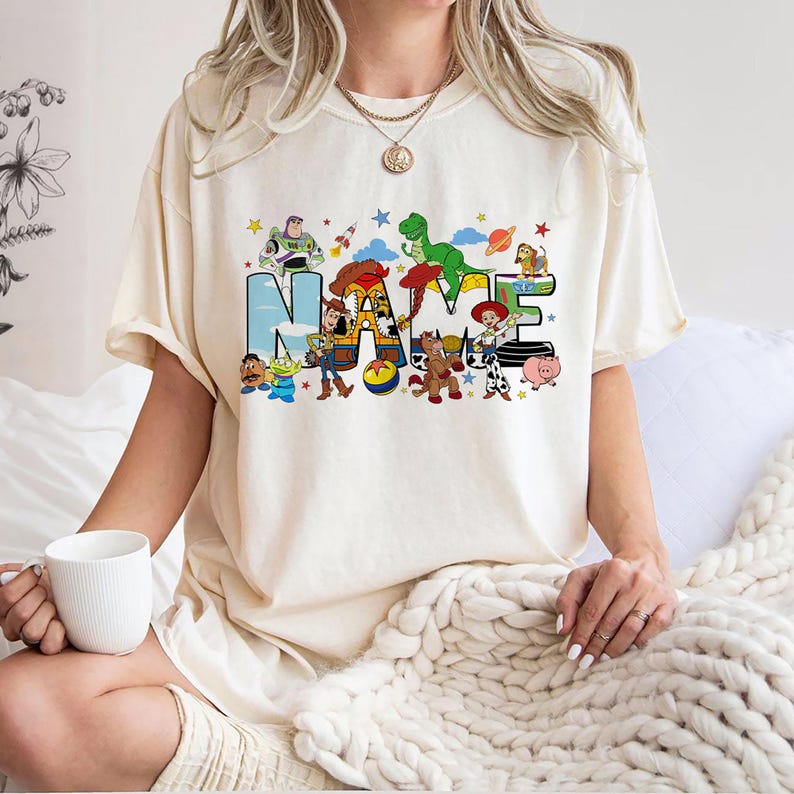 May include: Cream-colored t-shirt featuring the word "NAME" in a colorful, cartoonish font. The letters are decorated with characters from a popular animated movie. The shirt has short sleeves and a relaxed fit.