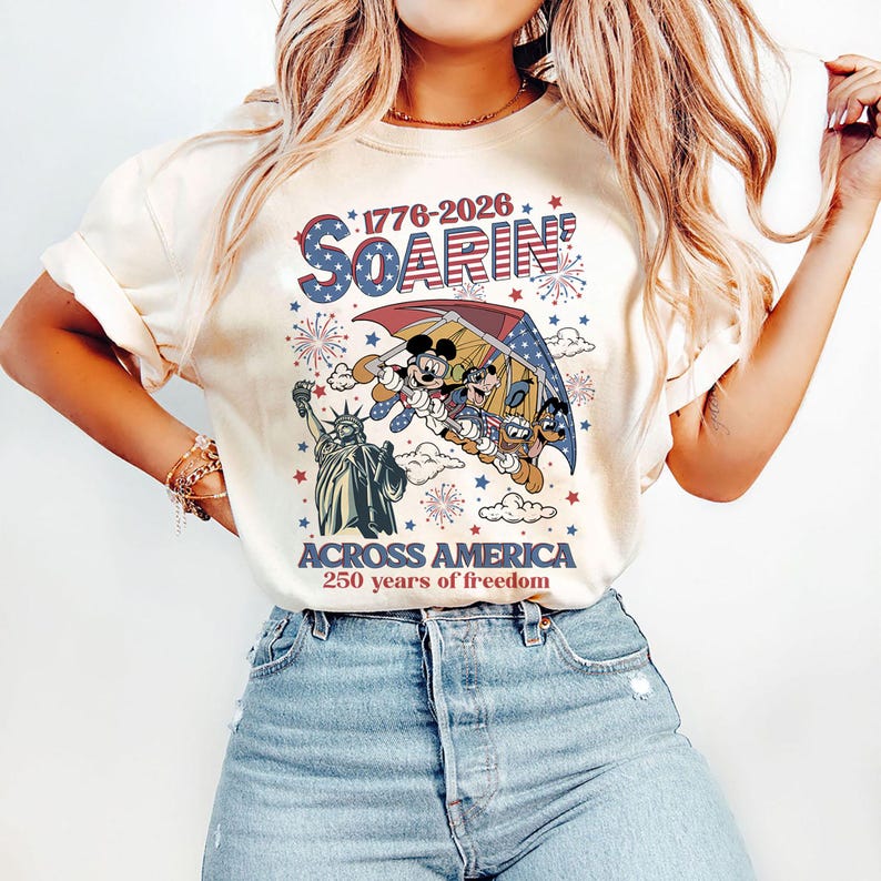 May include: Cream-colored t-shirt featuring a graphic of Mickey Mouse, Donald Duck, and Goofy soaring over the Statue of Liberty. The shirt has the text "1776-2026 SOARIN' ACROSS AMERICA 250 years of freedom" in red, white, and blue.