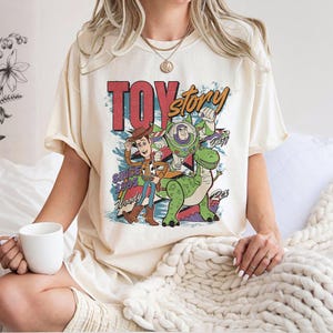 May include: Cream-colored t-shirt featuring a colorful graphic of Toy Story characters Woody, Buzz Lightyear, and Rex. The design includes the text "TOY STORY" in red and yellow, with "SINCE 1995" and character names. The shirt has short sleeves and a relaxed fit.