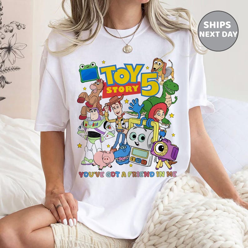 May include: A white t-shirt featuring a colorful graphic print of various animated characters from the Toy Story franchise, including Woody, Buzz Lightyear, and Rex. The text reads Toy Story 5 and You've Got A Friend In Me in multi-colored lettering.