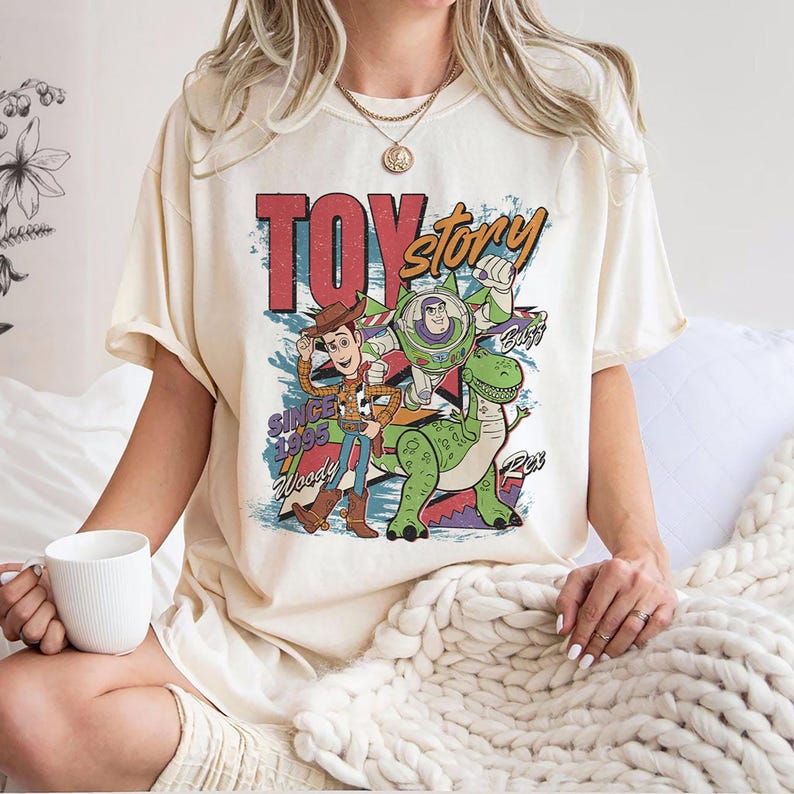 May include: Cream-colored t-shirt featuring a colorful graphic of Toy Story characters Woody, Buzz Lightyear, and Rex. The design includes the text "TOY STORY" and "SINCE 1995". The shirt has short sleeves and a relaxed fit.