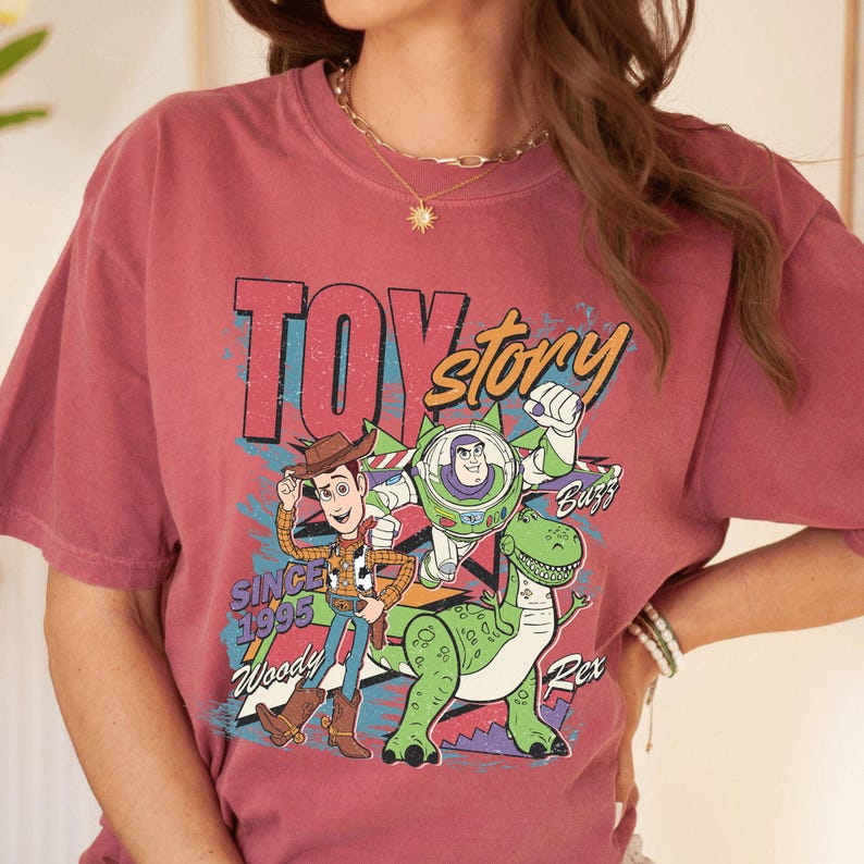 May include: A maroon t-shirt featuring a graphic of characters from the animated film "Toy Story." The design includes Woody, Buzz Lightyear, and Rex, with the text "TOY STORY" and "SINCE 1995." The shirt has short sleeves.