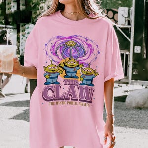 May include: A light pink t-shirt featuring a graphic of three green three-eyed aliens beneath a mechanical claw and a purple swirling portal. The text reads THE CLAW and THE MYSTIC PORTAL AWAITS in a bold purple font with a retro-inspired design.