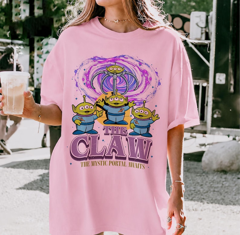 May include: A light pink t-shirt featuring a graphic of three green three-eyed aliens beneath a mechanical claw and a purple swirling portal. The text reads THE CLAW and THE MYSTIC PORTAL AWAITS in a bold purple font with a retro-inspired design.