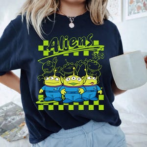 K&ouml;nnte beinhalten: Ein dunkelblaues T-Shirt mit einem leuchtend hellgr&uuml;nen Grafikdruck von drei drei&auml;ugigen Au&szlig;erirdischen in blauen Raumanz&uuml;gen. Das Design umfasst einen karierten Hintergrund, den Schriftzug "Aliens" oben und die Jahreszahl "1995" rechts.