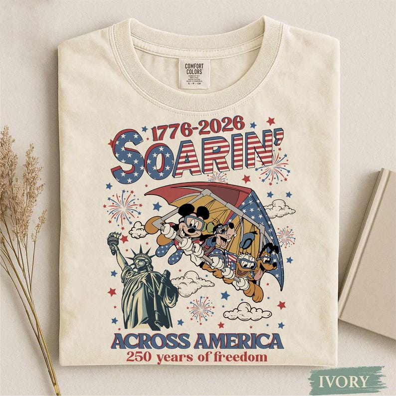 Może przedstawiać: Kremowa koszulka z patriotycznym wzorem. Przedstawia Myszka Miki, Goofy'ego i Pluto 'szybujących'. Tekst brzmi "1776-2026 Soarin' Across America 250 years of freedom" z czerwonymi, białymi i niebieskimi akcentami.