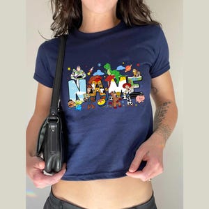 May include: Navy blue t-shirt featuring the word "NAME" in colorful letters, with cartoon characters from a popular animated movie. The shirt has short sleeves and a crew neck.