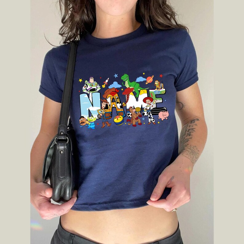 May include: Navy blue t-shirt featuring the word "NAME" in colorful letters, with cartoon characters from a popular animated movie. The shirt has short sleeves and a crew neck.