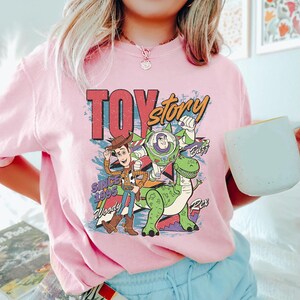 May include: Pink t-shirt featuring a colorful graphic of Toy Story characters Woody, Buzz Lightyear, and Rex. The design includes the text "TOY STORY" and "SINCE 1995". The shirt is a crew neck style.