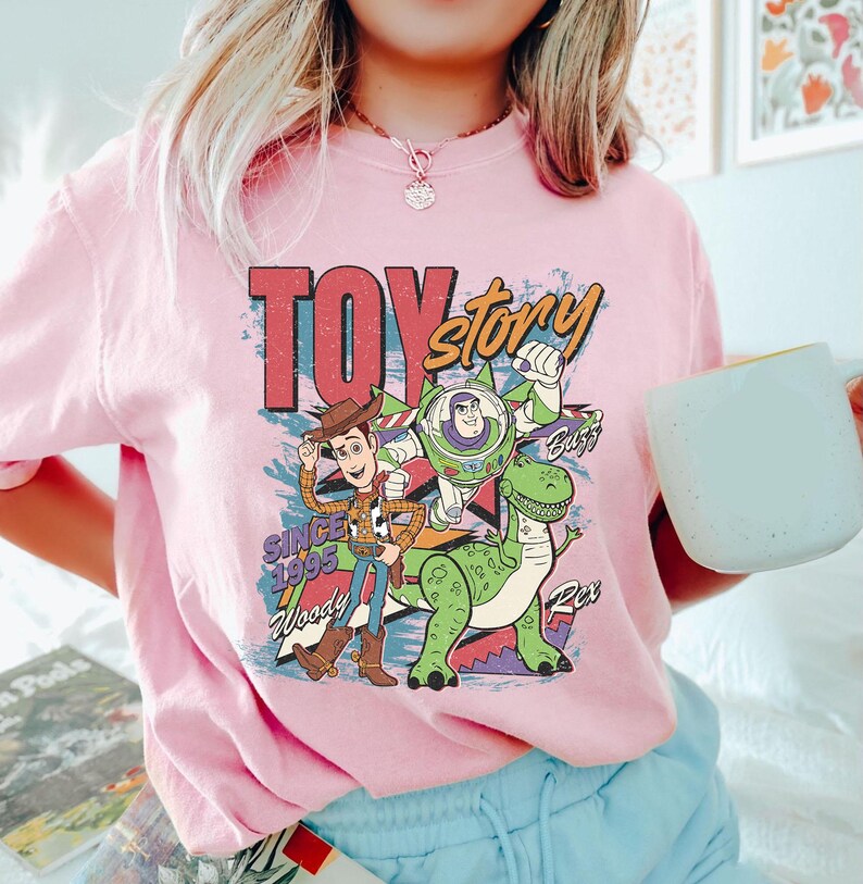 May include: Pink t-shirt featuring a colorful graphic of Toy Story characters Woody, Buzz Lightyear, and Rex. The design includes the text "TOY STORY" and "SINCE 1995". The shirt is a crew neck style.