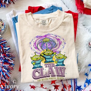 May include: A cream-colored t-shirt featuring a graphic of three green three-eyed aliens in blue space suits. Above them is a swirling purple and blue portal with a mechanical claw. The text reads THE CLAW THE MYSTIC PORTAL AWAITS.
