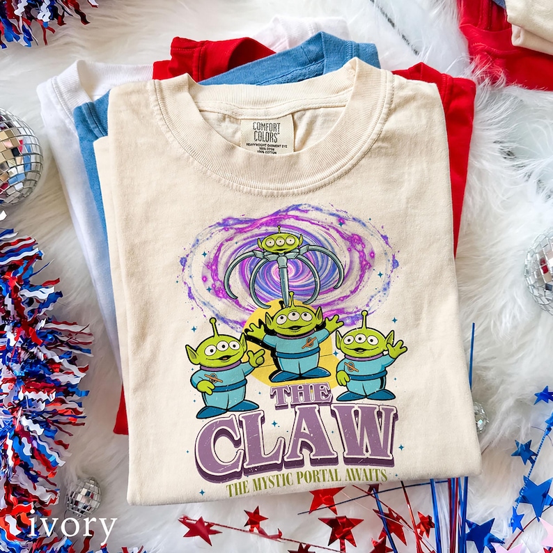May include: A cream-colored t-shirt featuring a graphic of three green three-eyed aliens in blue space suits. Above them is a swirling purple and blue portal with a mechanical claw. The text reads THE CLAW THE MYSTIC PORTAL AWAITS.