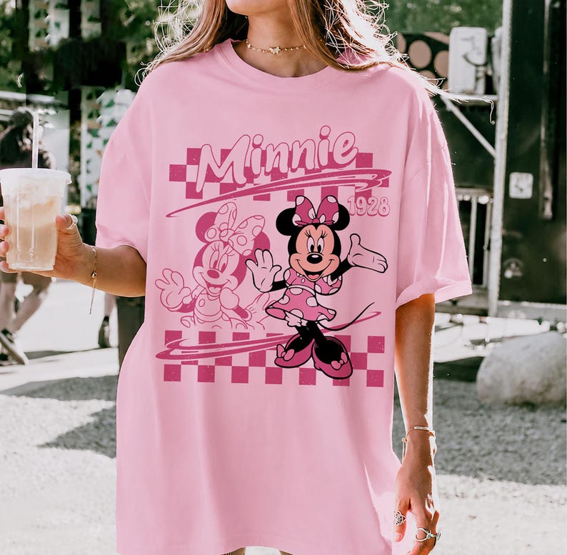 May include: A light pink t-shirt featuring a graphic print of Minnie Mouse. The design includes the text "Minnie" and "1928" over a pink and white checkered background, with a secondary outline illustration of the character positioned to the left.