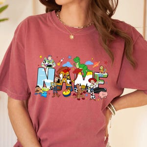 May include: A dusty rose t-shirt featuring the word "NAME" in colorful, cartoon-style letters. The letters are decorated with characters from a popular animated movie. Stars and other graphics are scattered around the letters.