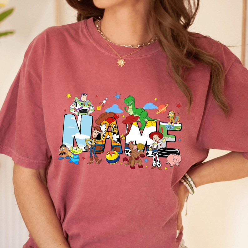 May include: A dusty rose t-shirt featuring the word "NAME" in colorful, cartoon-style letters. The letters are decorated with characters from a popular animated movie. Stars and other graphics are scattered around the letters.