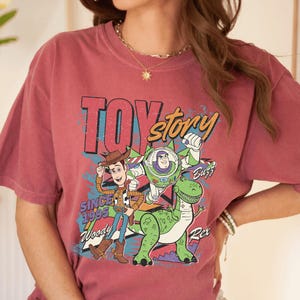 May include: A maroon-colored t-shirt featuring a graphic of characters from the animated film "Toy Story." The design includes Woody, Buzz Lightyear, and Rex, with the text "TOY STORY" and "SINCE 1995."