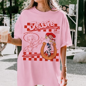 K&ouml;nnte beinhalten: Ein hellrosa T-Shirt mit kurzen &Auml;rmeln und einem rot-wei&szlig; karierten Hintergrundmuster. Das Design enth&auml;lt den Schriftzug Jessie 199s, eine Strichzeichnung der Figur Jessie und ein farbiges Bild von Jessie innerhalb eines Hufeisens.