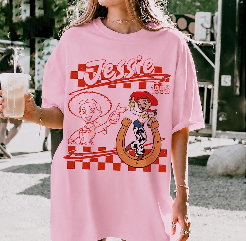 K&ouml;nnte beinhalten: Ein hellrosa T-Shirt mit kurzen &Auml;rmeln und einem rot-wei&szlig; karierten Hintergrundmuster. Das Design enth&auml;lt den Schriftzug Jessie 199s, eine Strichzeichnung der Figur Jessie und ein farbiges Bild von Jessie innerhalb eines Hufeisens.