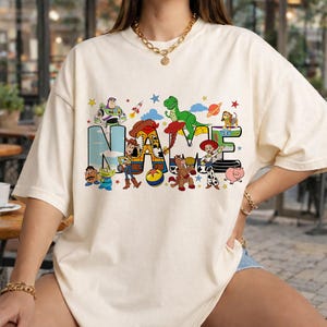 May include: Cream-colored t-shirt featuring the word "NAME" in a colorful, cartoon-style design. The letters are filled with images of characters from a popular animated movie. Stars and other decorative elements surround the word.