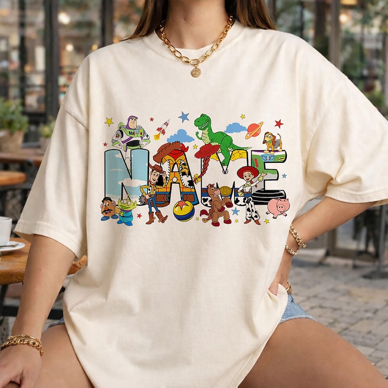 May include: Cream-colored t-shirt featuring the word "NAME" in a colorful, cartoon-style design. The letters are filled with images of characters from a popular animated movie. Stars and other decorative elements surround the word.