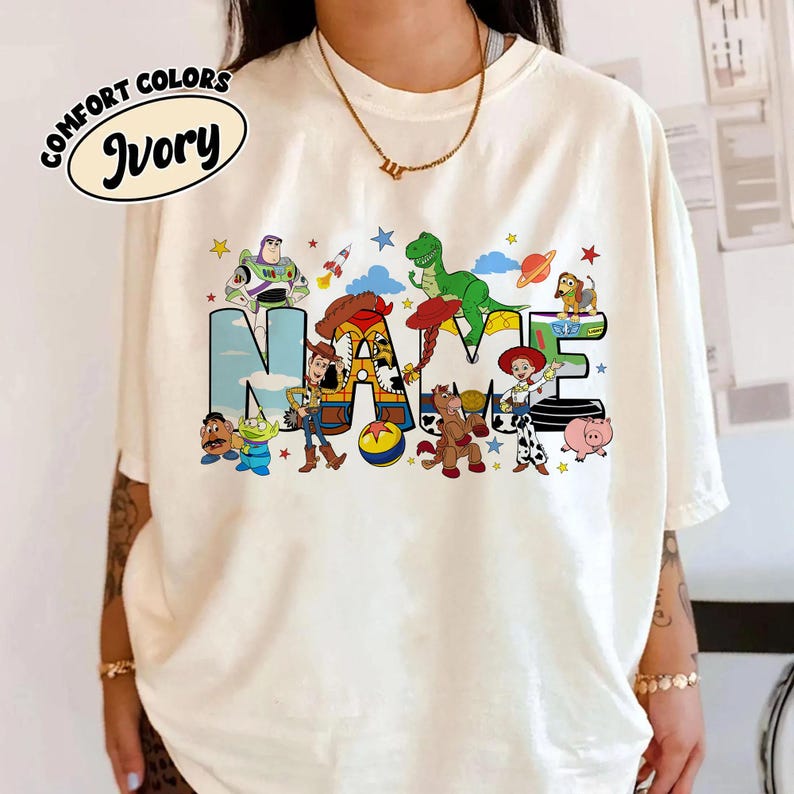 May include: Ivory-colored t-shirt with the text "NAME" in a colorful, cartoon-style design. The design features various characters and elements from a popular animated movie, including a green dinosaur, a space ship, and a cowboy.