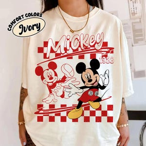 May include: An ivory t-shirt featuring a graphic print of Mickey Mouse in red and black. The design includes a red and white checkered pattern, the text Mickey, and the year 1928. A small oval label in the top left corner reads Comfort Colors Ivory.