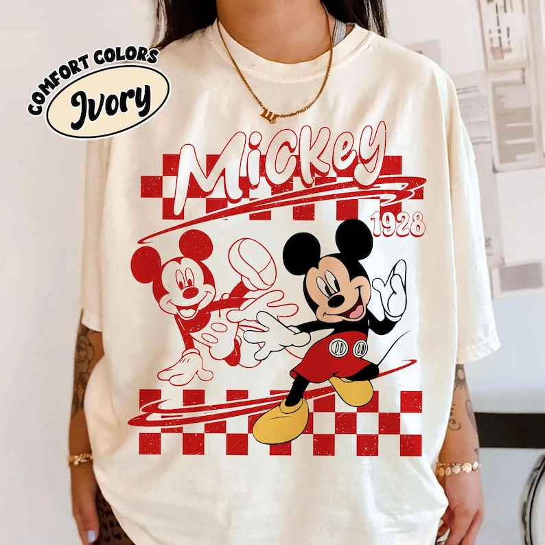 May include: An ivory t-shirt featuring a graphic print of Mickey Mouse in red and black. The design includes a red and white checkered pattern, the text Mickey, and the year 1928. A small oval label in the top left corner reads Comfort Colors Ivory.