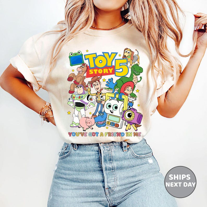May include: A cream-colored short-sleeve t-shirt featuring a colorful graphic print of Toy Story characters including Woody, Buzz Lightyear, Jessie, Rex, and Slinky Dog. The text reads Toy Story 5 and You've Got A Friend In Me with small yellow stars.