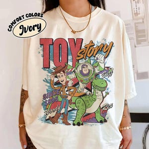 May include: Ivory-colored t-shirt featuring a graphic of Woody, Buzz Lightyear, and Rex from Toy Story. The design includes the words "TOY STORY" in red and orange, with "Since 1995" and character names. The shirt has short sleeves.