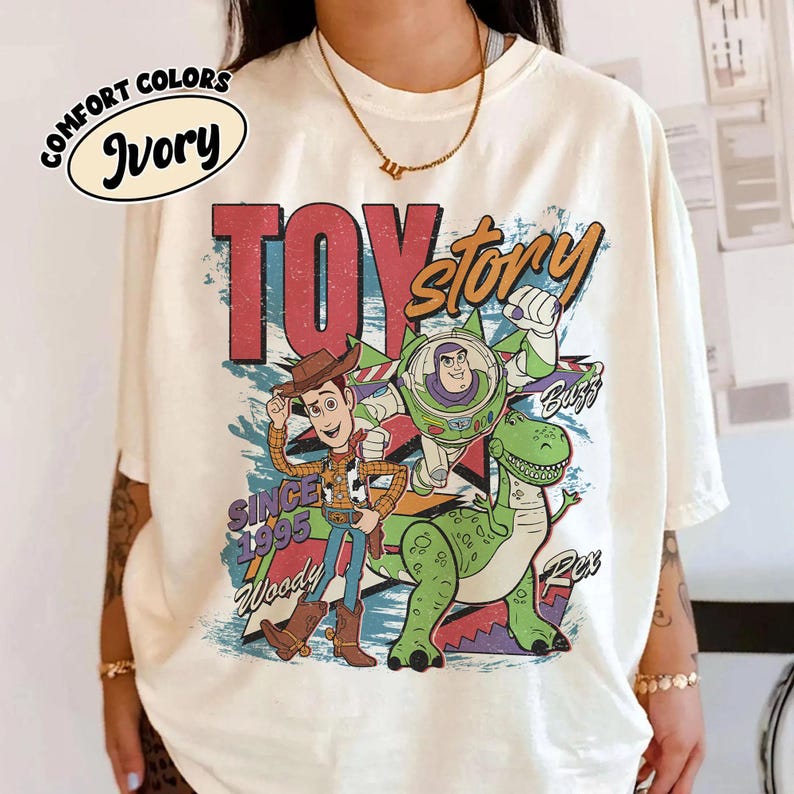 May include: Ivory-colored t-shirt featuring a graphic of Woody, Buzz Lightyear, and Rex from Toy Story. The design includes the words "TOY STORY" in red and orange, with "Since 1995" and character names. The shirt has short sleeves.