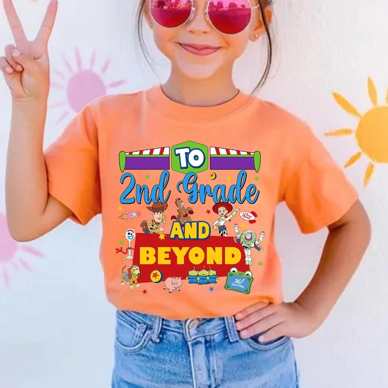 May include: An orange short-sleeved t-shirt featuring a graphic print with the text To 2nd Grade AND BEYOND. The design includes colorful illustrations of various animated toy characters, including a cowboy, a space ranger, and a cowgirl.