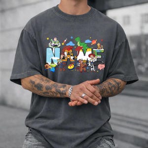 May include: Dark gray t-shirt with the word "NAME" in colorful, cartoon-style letters. The letters are decorated with various animated characters. The shirt is a relaxed fit.