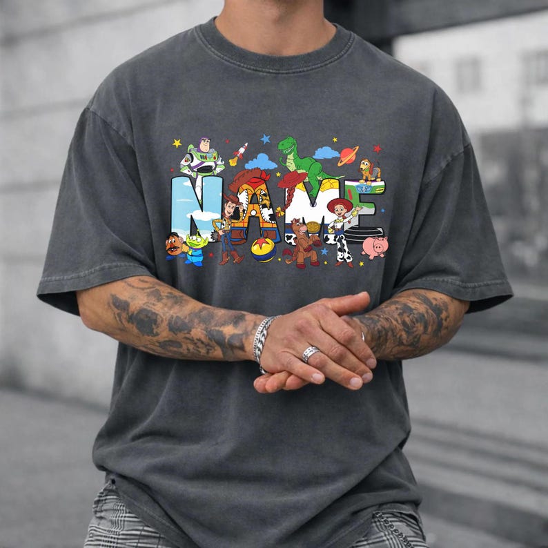 May include: Dark gray t-shirt with the word "NAME" in colorful, cartoon-style letters. The letters are decorated with various animated characters. The shirt is a relaxed fit.