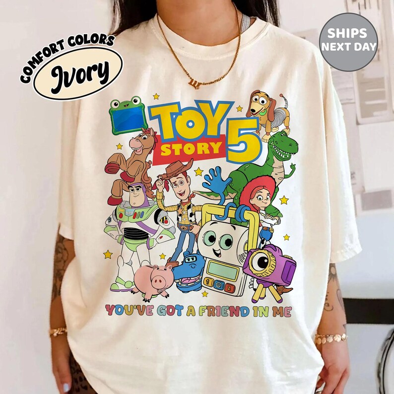 May include: An ivory t-shirt featuring a colorful graphic print of Toy Story characters including Woody, Buzz Lightyear, Rex, Slinky Dog, and others. The text reads "TOY STORY 5" and "YOU'VE GOT A FRIEND IN ME" in bold, multi-colored lettering across the front.