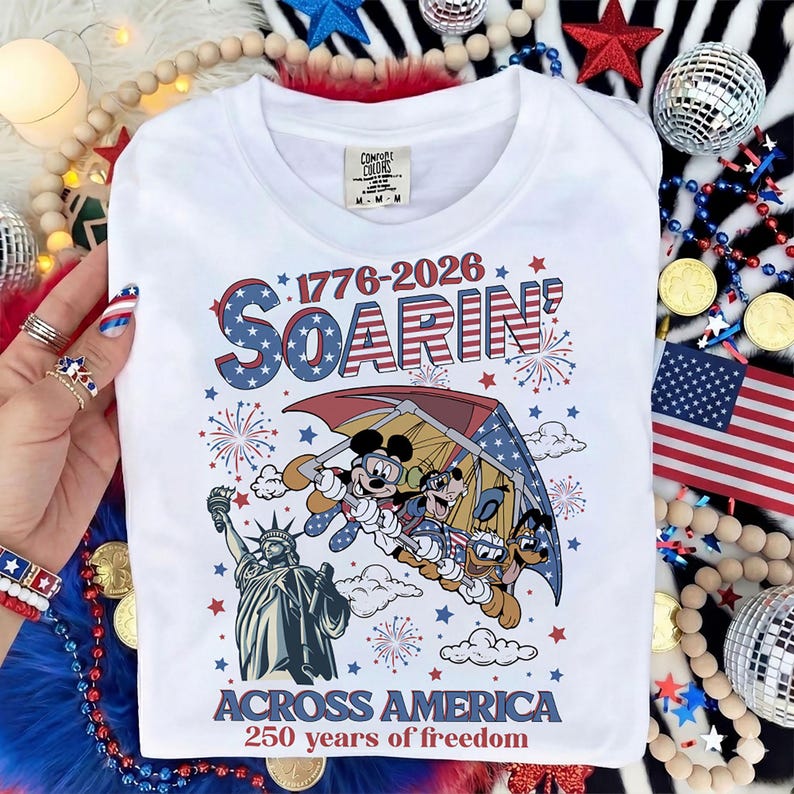 Może przedstawiać: Biała koszulka z patriotycznym wzorem przedstawiającym Myszka Miki, Kaczora Donalda i Goofy'ego szybujących nad Statuą Wolności. Koszulka ma napis "1776-2026 Soarin' Across America 250 years of freedom."