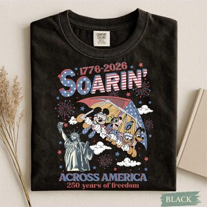 Może przedstawiać: Czarny t-shirt z grafiką Myszki Miki, Kaczora Donalda i Goofy'ego szybujących na paralotni nad Statuą Wolności. Koszulka zawiera napis "1776-2026 Soarin' Across America 250 years of freedom" w kolorach czerwonym, białym i niebieskim.